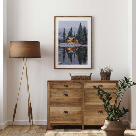 Christmas cabin photography print showing a warmly lit lodge beside a snowy lake under a pale winter sky.