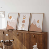 Boho wall art trio featuring an ochre sun, stacked moon phases and terracotta mountains on pale neutral backgrounds.