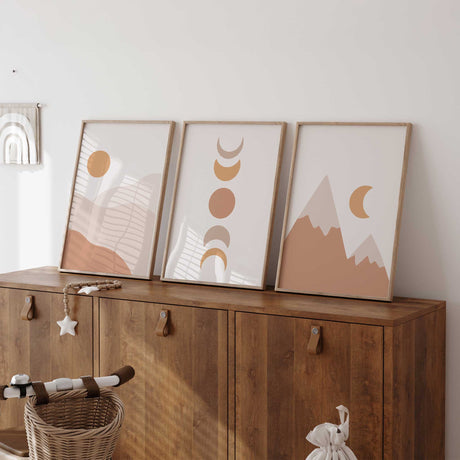 Boho wall art trio featuring an ochre sun, stacked moon phases and terracotta mountains on pale neutral backgrounds.