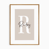 Personalised name print with a large white initial and black script lettering on a warm pink background.