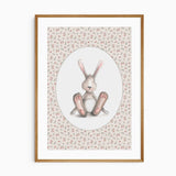Seated grey-brown bunny with pink ears and paws inside a white oval, surrounded by a pink rose and green leaf pattern.