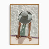 Overhead poolside photograph of a woman wearing a wide-brimmed sunhat in pale water, hands resting on the pool edge.