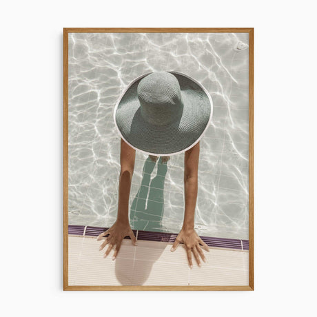 Overhead poolside photograph of a woman wearing a wide-brimmed sunhat in pale water, hands resting on the pool edge.