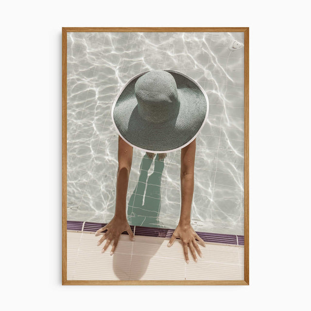 Overhead poolside photograph of a woman wearing a wide-brimmed sunhat in pale water, hands resting on the pool edge.