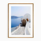 A framed art print depicting a scenic view of Santorini with white buildings, a cobblestone path, and a deep blue sea under a clear sky.