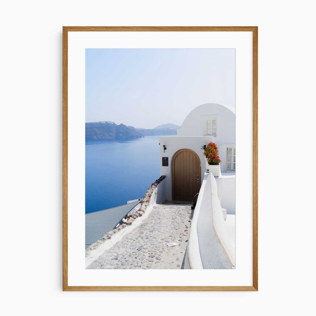 A framed art print depicting a scenic view of Santorini with white buildings, a cobblestone path, and a deep blue sea under a clear sky.