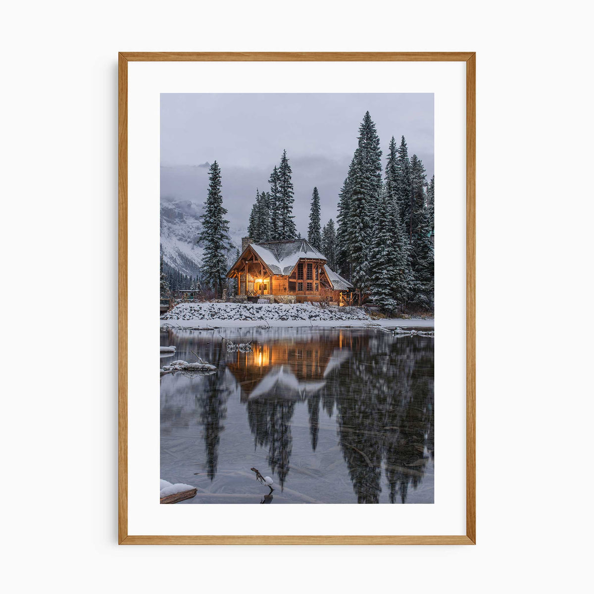 Photographic print of a glowing wooden cabin beside a snowy lake, framed by pine trees and a clean white border.