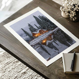 Unframed snowy cabin photo print with an even white border around the lake, forest and glowing wooden cabin.