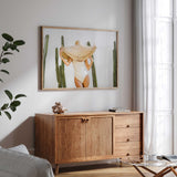 Woman in a straw sunhat among green cacti, shown as a landscape wall art print above a timber and cane sideboard.